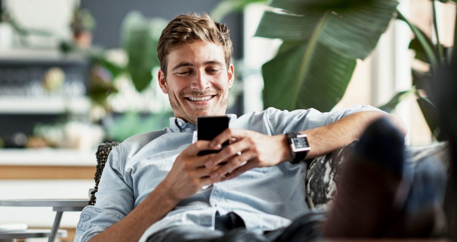 Mann liegt lächelnd auf der Couch mit seinem Handy in der Hand
